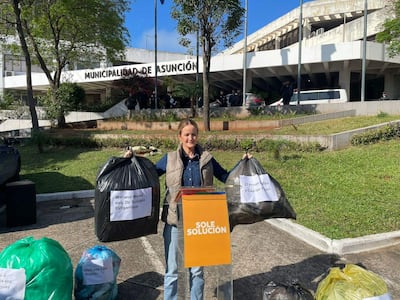 Soledad Núñez, con camisa de mezclilla y jeans, sostiene bolsas de basura frente a la Municipalidad de Asunción, rodeada de hombres de seguridad.