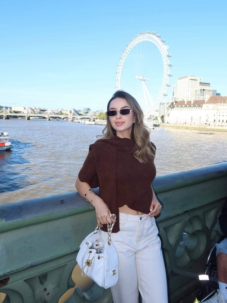 Mujer de pie con top marrón y pantalones blancos, sosteniendo un bolso blanco, junto al río con la London Eye detrás.
