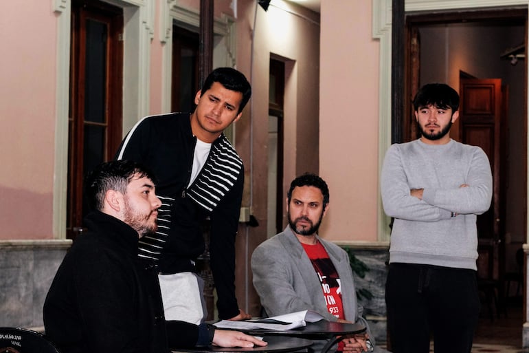 Hombres en ambiente formal discuten; uno con abrigo negro habla, mientras otros escuchan atentos, vestidos de forma casual y formal.