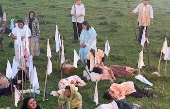 Dos mujeres, una abrazando a un niño, en un campo, vestidas con ropas antiguas y manchadas de sangre, rodeadas de personas con antorchas.