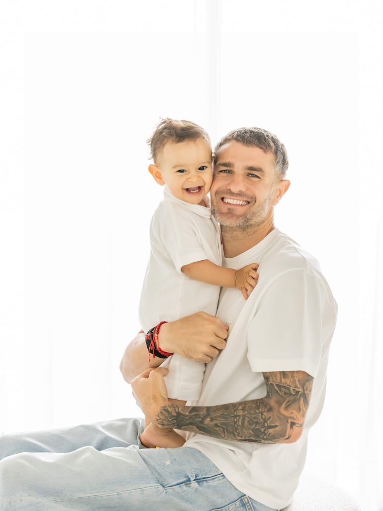 Pablo Vegetti con cabello corto y tatuajes sonríe mientras sostiene a un niño de cabello rizado que viste una camisa blanca.