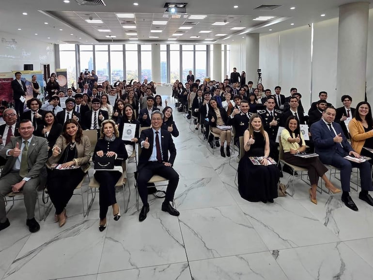 Tres jóvenes levantan el pulgar en señal de celebración, rodeados por una multitud en trajes formales en un aula iluminada.