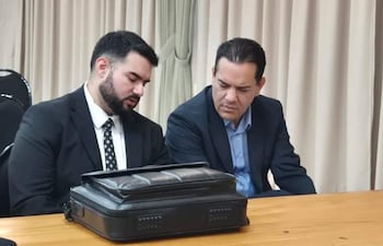 Friedmann Alfaro con traje, barba y cabello corto, discutiendo con un hombre de cabello liso en un entorno formal.