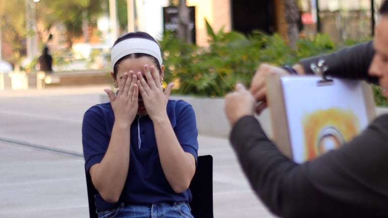 Niño con camiseta azul y diadema blanca cubriendo su rostro, mientras un adulto observa un dibujo en un caballete.