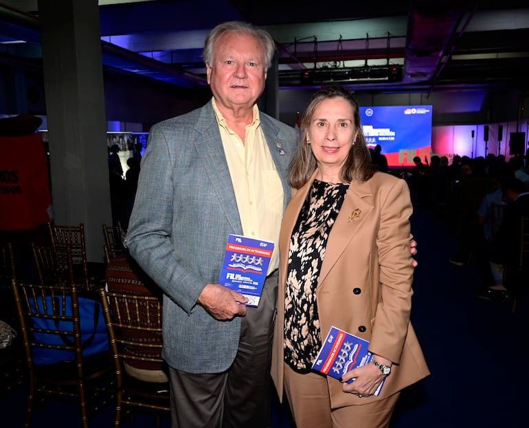 Peter Hansen, con saco gris y camisa amarilla, sostiene un folleto; la mujer, en blusa estampada y abrigo claro, sonríe a la cámara.