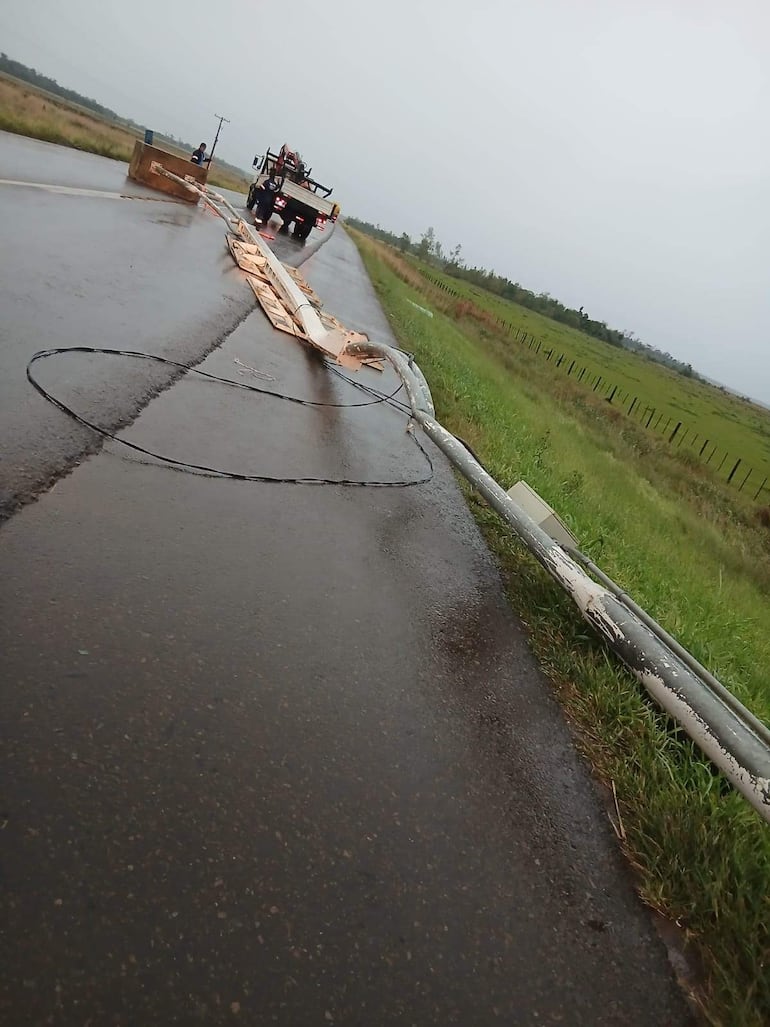 Dos personas observan un camión retirando un poste caído sobre la carretera en un día lluvioso con charcos en el asfalto.