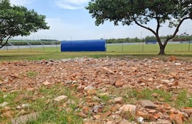 Terreno con escombros y dos árboles, campo de fútbol al fondo con gradas y refugio azul.