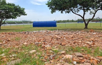 Terreno con escombros y dos árboles, campo de fútbol al fondo con gradas y refugio azul.