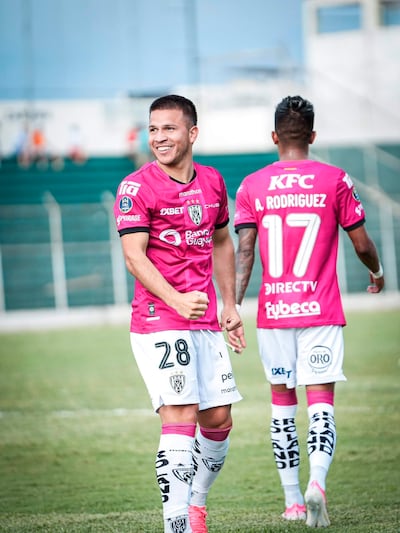Hugo Quintana sonriente con camiseta rosa y número 28, mientras otro jugador relaxa a su lado en un campo de fútbol.