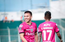 Hugo Quintana sonriente con camiseta rosa y número 28, mientras otro jugador relaxa a su lado en un campo de fútbol.