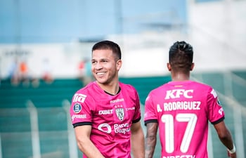 Hugo Quintana sonriente con camiseta rosa y número 28, mientras otro jugador relaxa a su lado en un campo de fútbol.