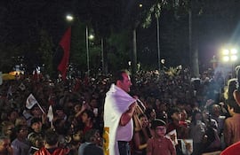 Hombre con camiseta blanca y bandera sobre los hombros, rodeado de una multitud efervescente con pancartas rojas y negras.