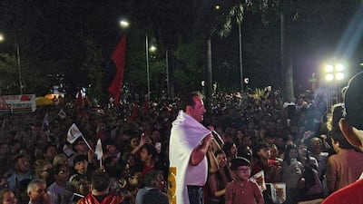 Hombre con camiseta blanca y bandera sobre los hombros, rodeado de una multitud efervescente con pancartas rojas y negras.
