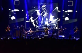Andrés Calamaro en el centro del escenario, tocando piano rodeado de músicos, mientras luces brillantes iluminan el ambiente oscuro.