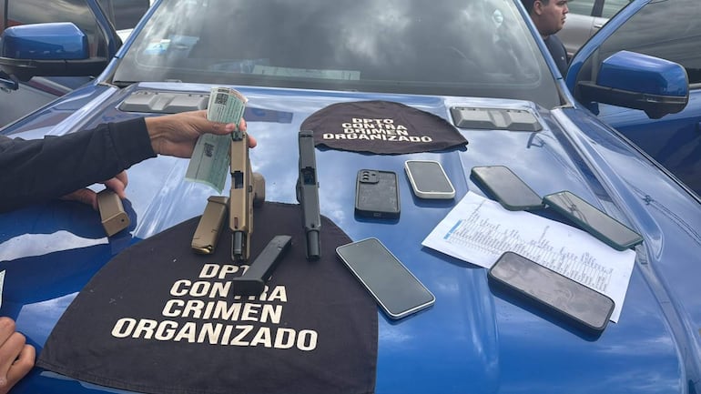 Armas semi-automáticas y teléfonos móviles sobre el capó de una camioneta, con documentos visibles y el paño del departamento de policía.