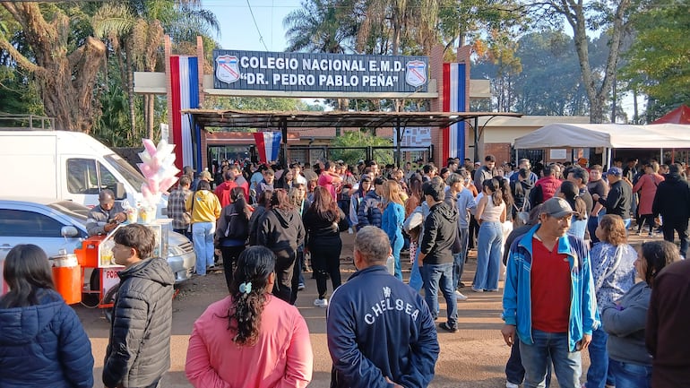 Multitud de personas frente al colegio, algunos en chaquetas de fútbol, socializando y comprando alimentos. Ambiente festivo.