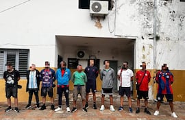 Ocho personas en exterior, usando camisetas de fútbol y pantalones cortos, parecen esperar un evento deportivo en un ambiente desgastado.