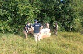 Dos agentes de policía rodean un auto semienterrado en vegetación, concentrados en la inspección mientras otros observan el entorno.