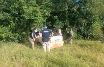 Dos agentes de policía rodean un auto semienterrado en vegetación, concentrados en la inspección mientras otros observan el entorno.