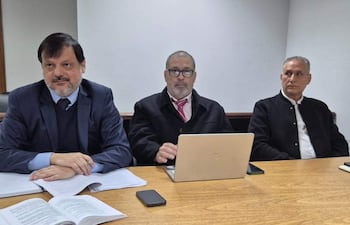 Claudio Lovera en traje oscuro habla, mientras Mario Elizeche escucha atentamente en camisa clara y corbata. Óscar Boidanich observa desde la derecha.