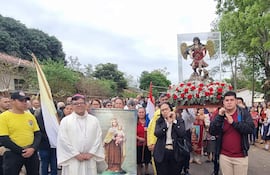 Sacerdote en túnica blanca sostiene imagen de la Virgen del Carmen, mientras fieles con camisetas amarillas lo siguen en una ceremonia solemne.