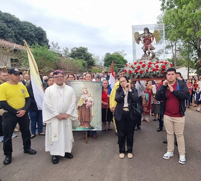 Sacerdote en túnica blanca sostiene imagen de la Virgen del Carmen, mientras fieles con camisetas amarillas lo siguen en una ceremonia solemne.