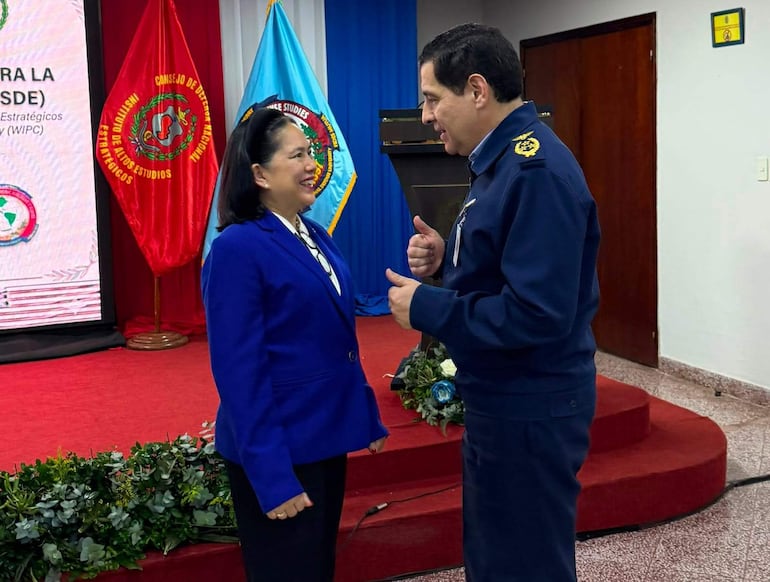 Celia Realuyo en saco azul y blusa blanca dialoga con Gral. Edwin Martinez en uniforme azul, en un entorno formal decorado con banderas.