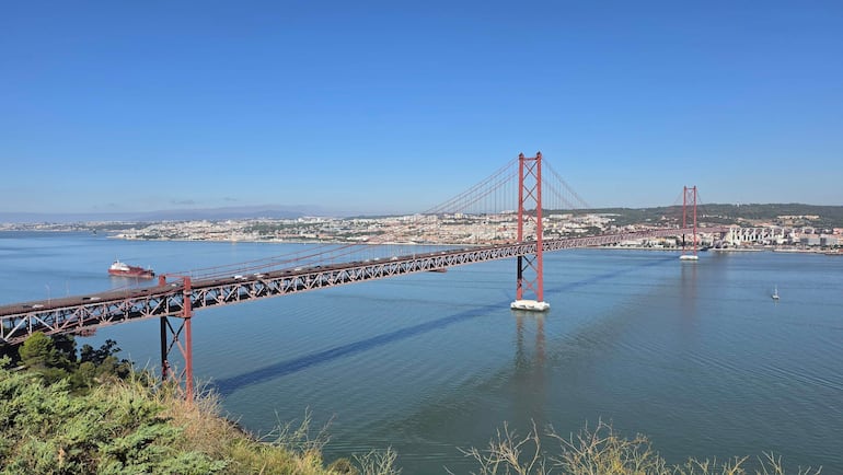 Puente 25 de Abril de color rojo sobre el río Tajo, con un entorno urbano y cielo despejado en Lisboa.