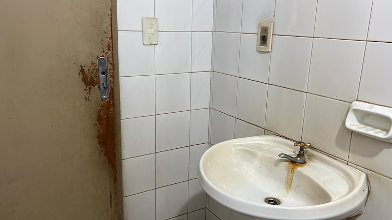 El sanitario de damas y caballeros presenta deterioro general: puerta dañada y sin picaporte, canillas oxidadas y mal olor en el ambiente, reflejando falta de mantenimiento.