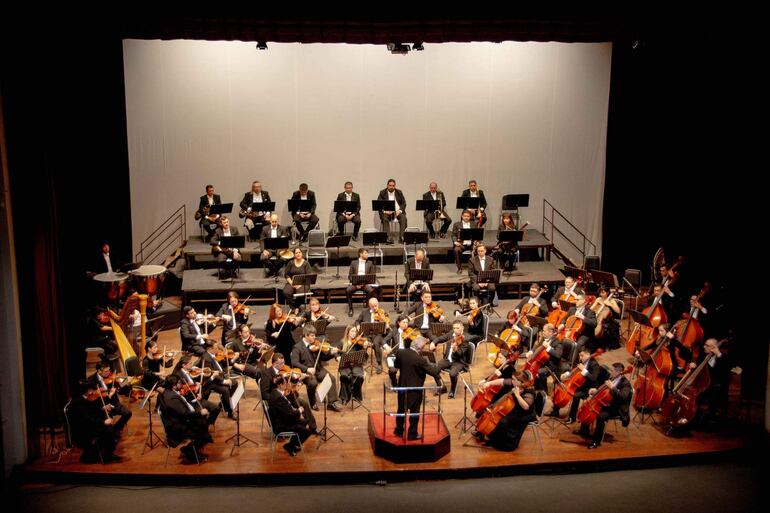 Orquesta con 40 músicos en trajes oscuros, director al frente dirigiendo con atención.