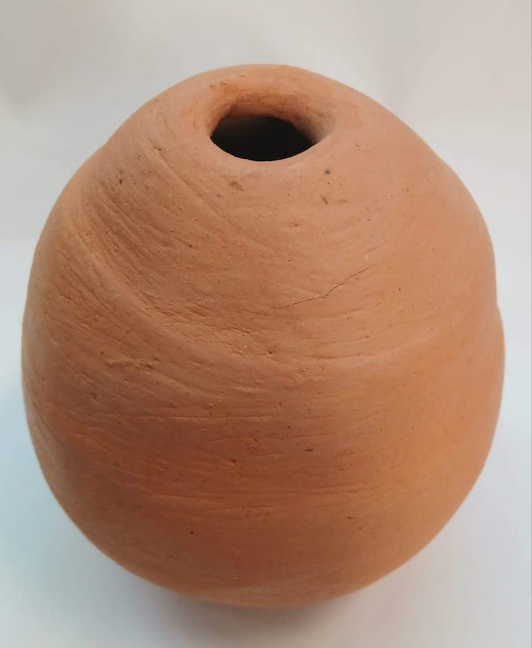 Jarra de barro terracota, de forma esférica y superficie lisa con algunas marcas, sobre fondo blanco.