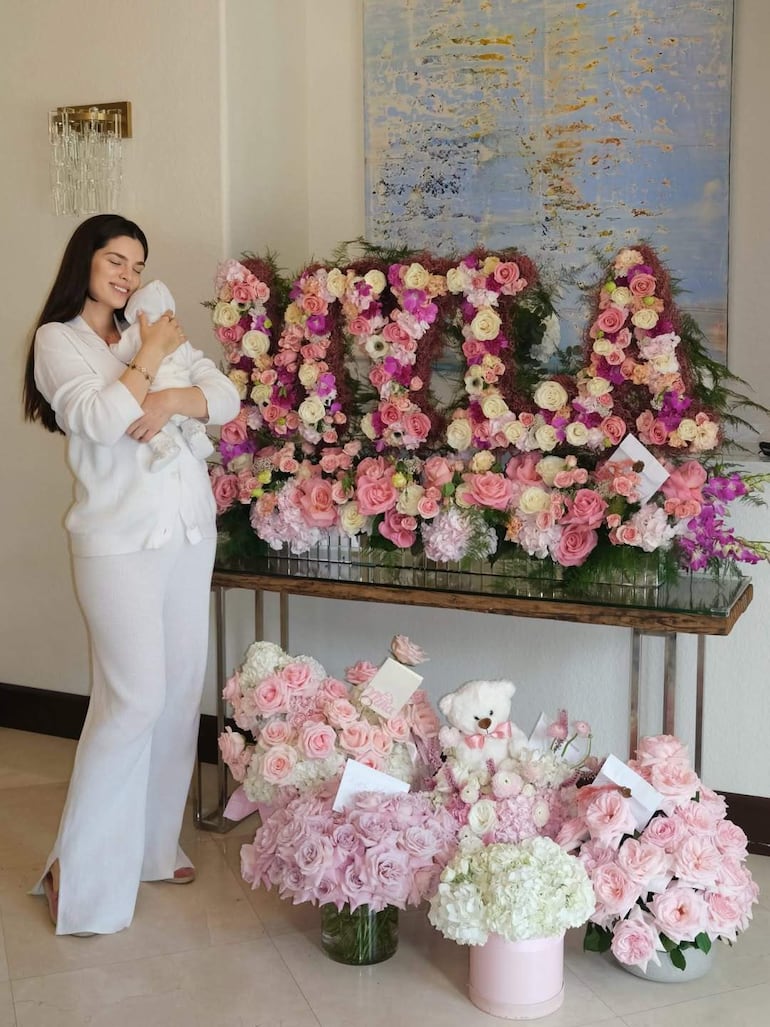 Mujer con expresión feliz sostiene un bebé envuelto, rodeada de flores rosas y blancas en un ambiente festivo.