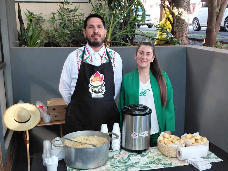 Hombre de barba y delantal negro y mujer con abrigo verde colaboran en puesto gastronómico del festival con comida y sombrero de paja.