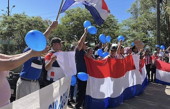 Asistentes en acto cívico al aire libre sosteniendo globos azules y banderas con colores de la patria, expresando entusiasmo.
