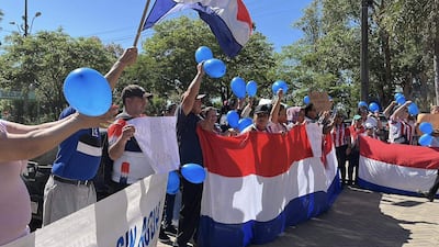 Asistentes en acto cívico al aire libre sosteniendo globos azules y banderas con colores de la patria, expresando entusiasmo.
