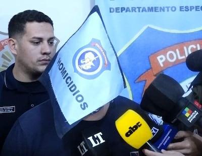 Hombre con gorra negra y camiseta sostiene bandera 'HOMICIDIOS', mientras un policía de uniforme observa en el fondo.