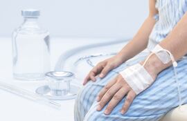 Manos de un hombre con bata de hospital y un suero intravenoso sentado en una cama.