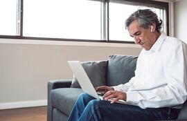 Hombre adulto con auriculares inalámbricos trabajando concentrado en su laptop desde el sofá de casa. La imagen transmite productividad, flexibilidad y comodidad, ideal para contenido sobre teletrabajo, freelancers, tecnología para adultos o estilos de vida modernos y conectados. Muestra un ambiente hogareño con buena luz natural.