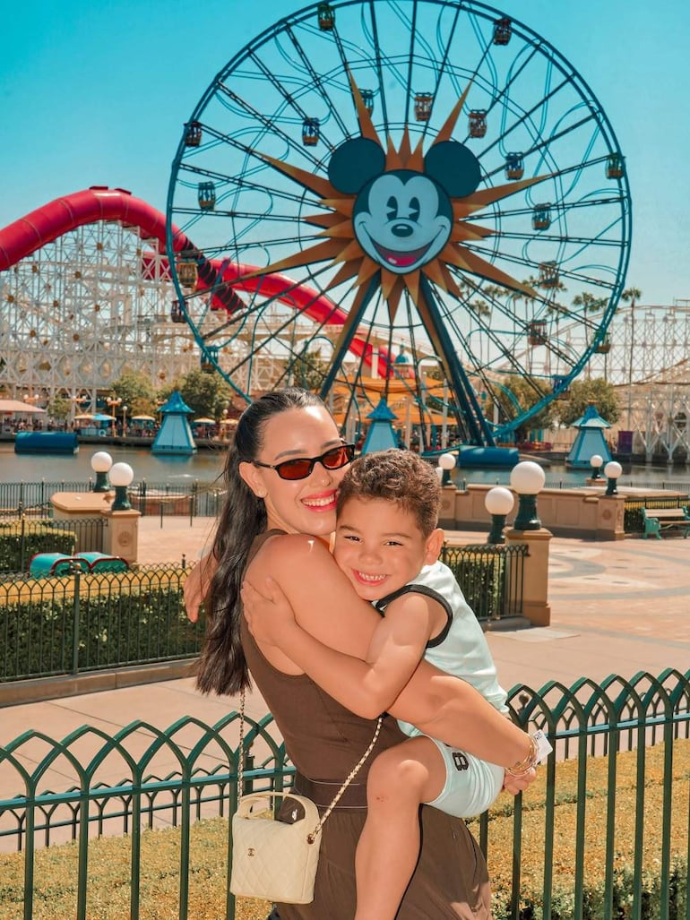 Mujer con gafas de sol y vestido marrón sostiene a un niño feliz, frente a una rueda de la fortuna en un parque de diversiones.