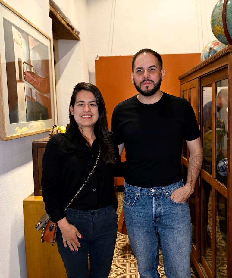 Pareja sonriente en una galería; la mujer viste camisa y pantalones negros, el hombre lleva camiseta negra y jeans claros.
