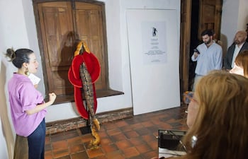 Paz Moreno Re con camisa lila, hablando con entusiasmo, mientras personas observan en un espacio expositivo.