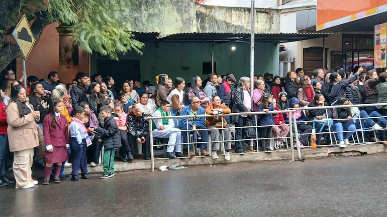 Grupo diverso de personas observando el desfile, algunos con teléfonos móviles, en un ambiente urbano.