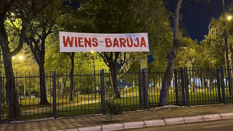 Cartel grande con 'WIENS-BARUJA' en letras rojas sobre fondo blanco, en un espacio público con árboles y área verde.