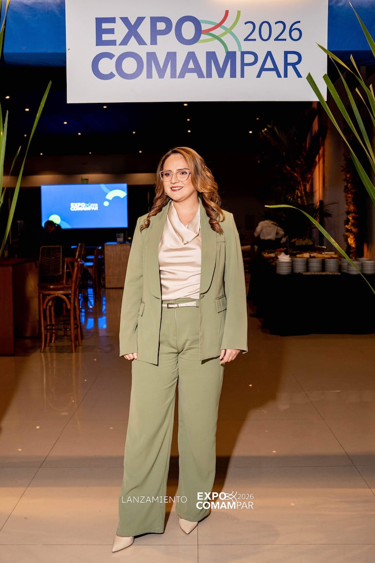 Mujer en traje verde claro y blusa beige sonríe frente a cartel de la Expo 2026 en un salón decorado.