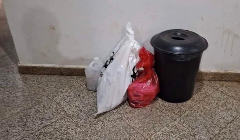 Un bote de basura negro y tres bolsas en el suelo de un pasillo del hospital, con paredes manchadas.