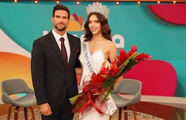 Gretha Matiauda con corona y vestido blanco sonríe sosteniendo un ramo, junto a un hombre en traje oscuro en un ambiente colorido.