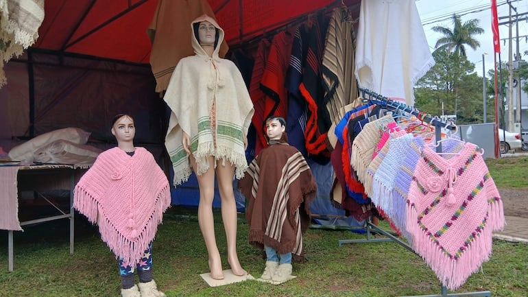 Tres maniquíes exhiben ponchos de diferentes colores en una feria artesanal, rodeados de otros productos textiles.