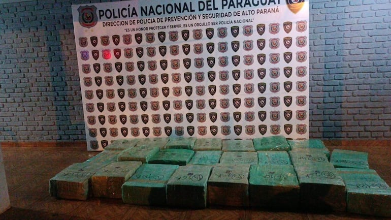 Panel de la Policía Nacional con escudos, frente a paquetes de marihuana envueltos en plástico verde.