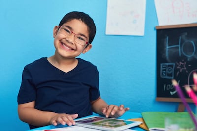 La imagen muestra a un niño sentado en un entorno que parece ser una habitación o un estudio. El niño tiene una sonrisa amplia y está usando gafas. Lleva una camiseta de color oscuro y está en el centro de la imagen, interactuando con una tablet que tiene en sus manos. Frente a él, hay elementos de papelería, incluidos cuadernos y un recipiente con lápices de colores.
Al fondo, hay una pared pintada de azul, decorada con dibujos de niños que están colgados. Adyacente, se encuentra una pizarra negra con palabras escritas en tiza. El ambiente es interior y parece estar diseñado para actividades escolares o creativas.
No hay personas adicionales visibles en la imagen. No se identifican elementos relacionados con fútbol, figuras públicas o lugares reconocibles. Las acciones que se están realizando parecen ser de estudio o recreativas.
