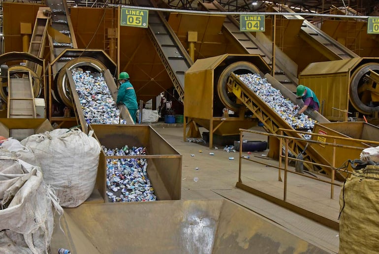 Dos trabajadores en overoles y cascos de seguridad operan cintas transportadoras con latas en un entorno industrial.
